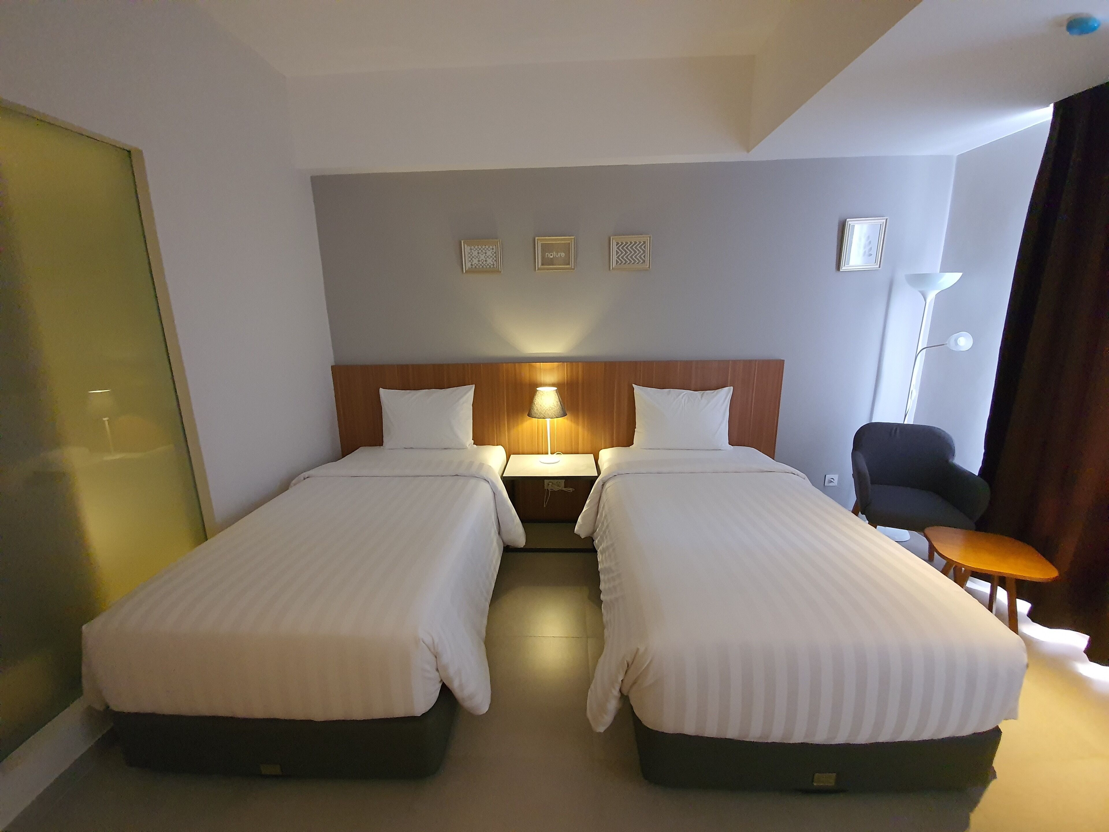 A hotel room with two single beds, a wooden headboard, a nightstand with a lamp, a chair, and a small table.