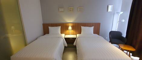 A hotel room with two single beds, a wooden headboard, a nightstand with a lamp, a chair, and a small table.