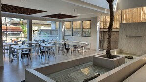 Un espace de restauration intérieur moderne, doté de tables et de chaises, d’un élément décoratif central avec de l’eau et de grandes fenêtres laissant entrer la lumière naturelle.