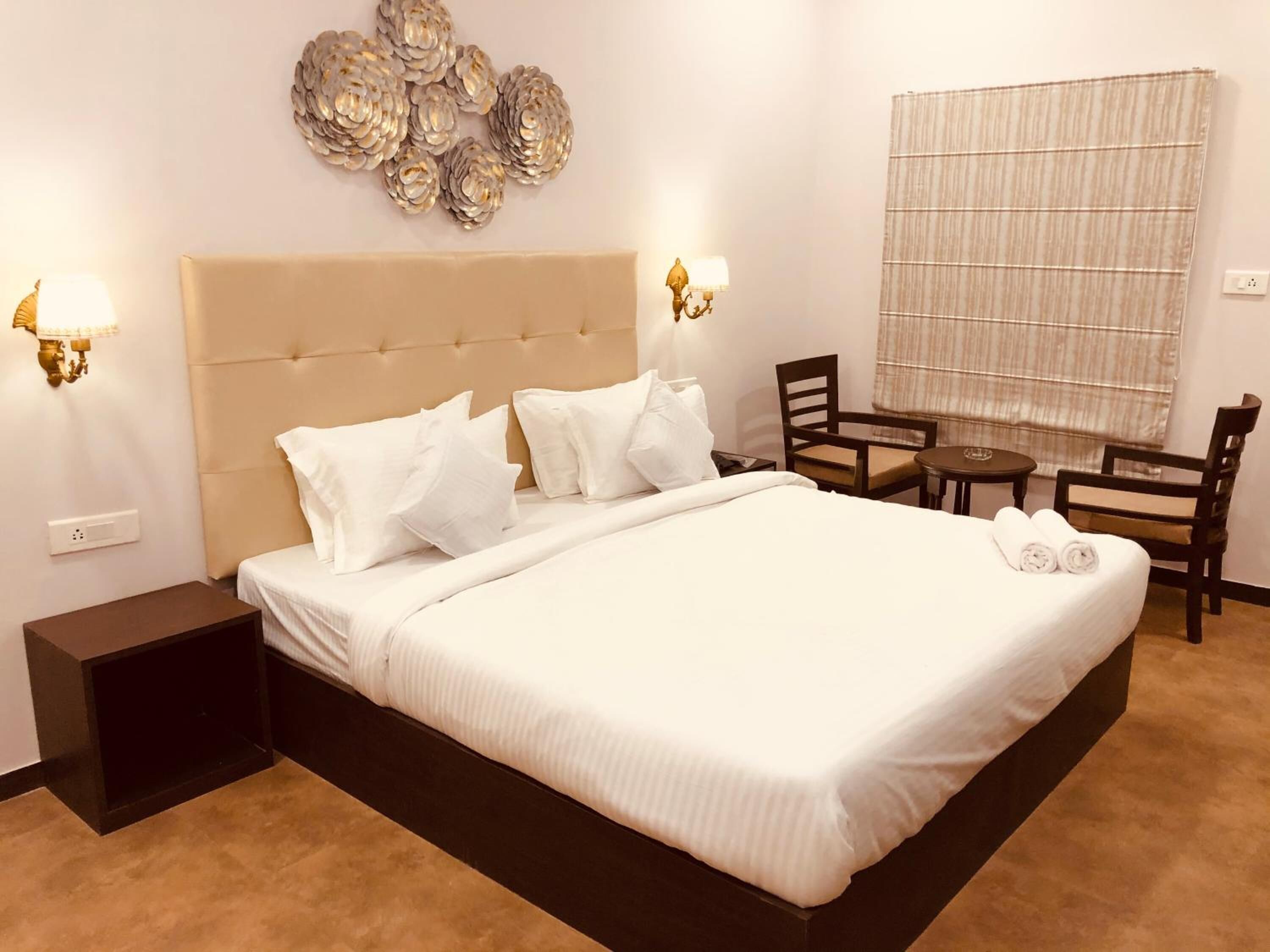 Habitación de hotel con una cama grande, dos mesitas de noche, una silla, una pequeña mesa y un adorno de pared.