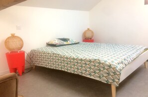 A small, simple room with a bed, a red stool, and a decorative vase.