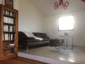 A modern living room with a dark grey sofa, a wooden bookshelf filled with books, a small table with a glass container, and a large window providing natural light.