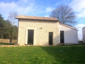 A stone building with two doors and a tiled roof, surrounded by a grassy yard and trees.