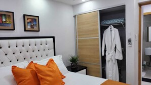 Ett hotellrum med en säng som har ett stoppat huvudgavel, två orange kuddar, en vit badrock som hänger på en klädställning och en liten krukväxt på ett sidobord.