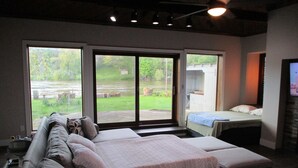 Et soveværelse med en stor seng, en sofa og et fjernsyn. Skydedøre fører ud til en terrasse med udsigt over sø og grøn bevoksning.