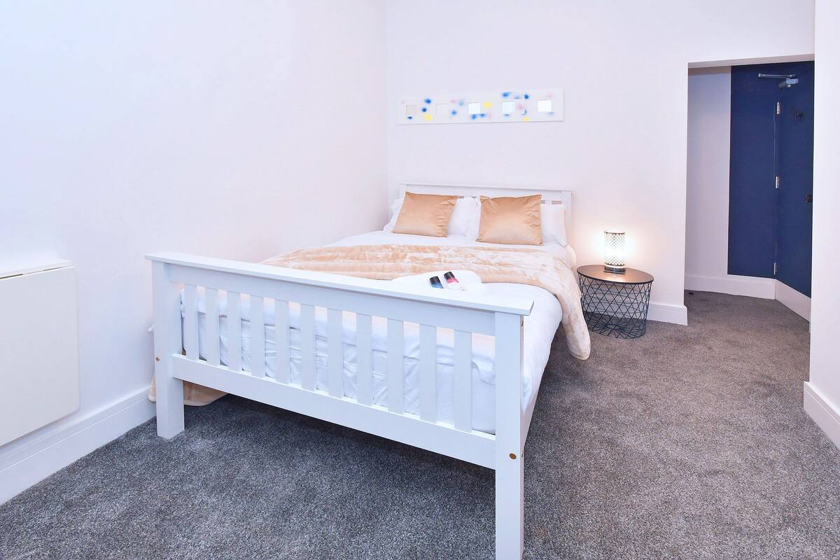 A bedroom with a white bed, a bedside table with a lamp, and a door leading to another room.