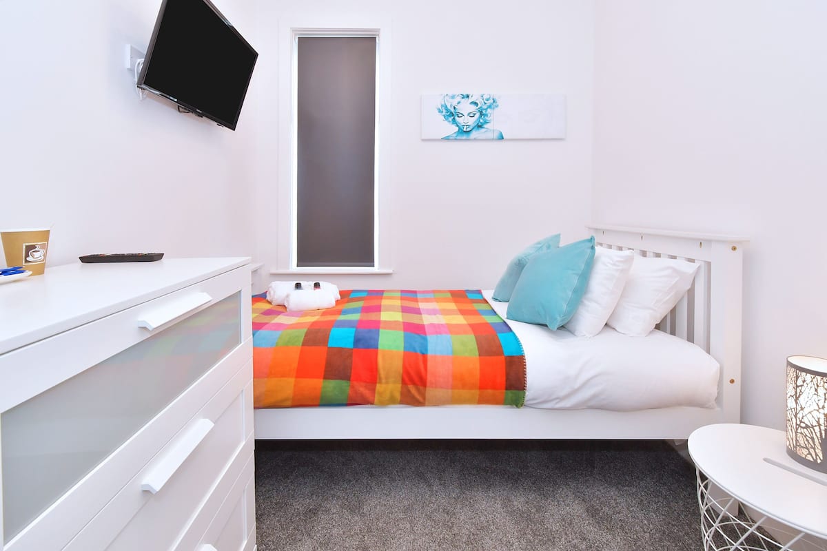 A bedroom with a bed, a desk, a chair, a TV, and a window.