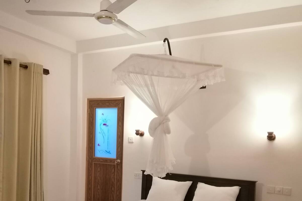 Quarto com cama, ventoinha no teto, porta e luminária fixada na parede.