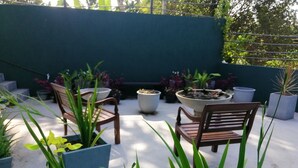 Un espace patio aménagé avec deux chaises en bois, des plantes en pot et un grand bac à fleurs agrémenté d’un élément décoratif aquatique.