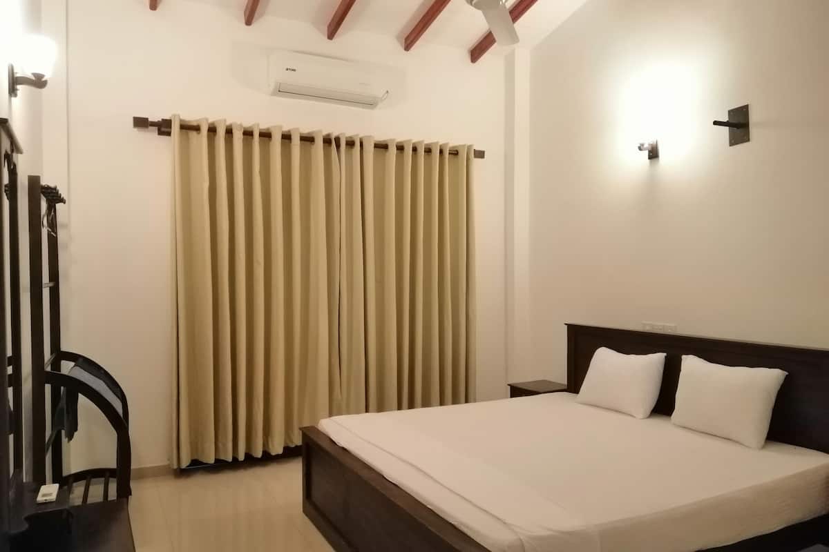 Quarto de hotel com cama, mesa de cabeceira, escrivaninha, cadeira, janela com cortinas, ar condicionado e ventilador de teto.