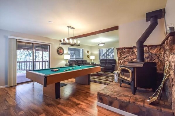 A spacious living room with a pool table, comfortable seating, a fireplace, and a view through a large window.