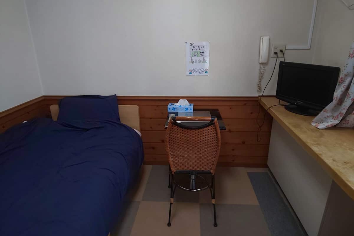 A small room with a bed, a desk, a chair, a television, and a wall-mounted light switch.
