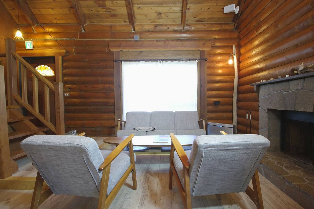 A log cabin interior with a wooden staircase, a sofa, two armchairs, a table, and a fireplace.