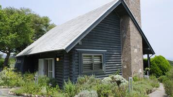 Une cabane en rondins avec une cheminée en pierre, entourée d’une variété de plantes et d’arbustes.