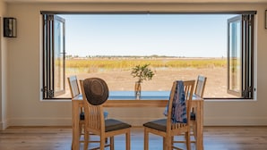 A room with a large window overlooking a field. There are two wooden chairs and a table with a hat and a plant on it. - L'Ermitage Quagga Lodge (Bergrivier)