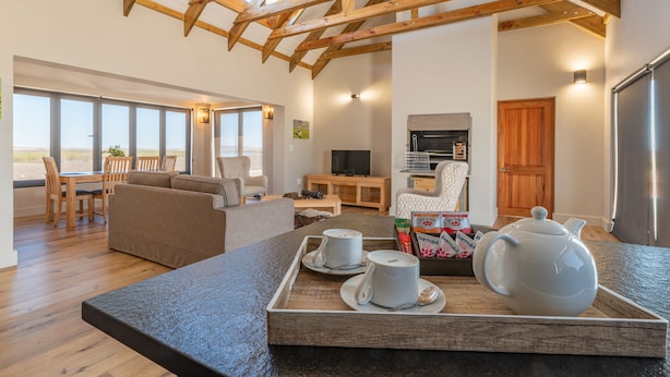 A spacious living area with a high ceiling, wooden beams, a dining table with chairs, a sofa, a television, and a kitchen area with a refrigerator. - L'Ermitage Quagga Lodge (Bergrivier)