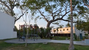 A playground with swings and climbing structures, surrounded by trees and a building with a white facade.
