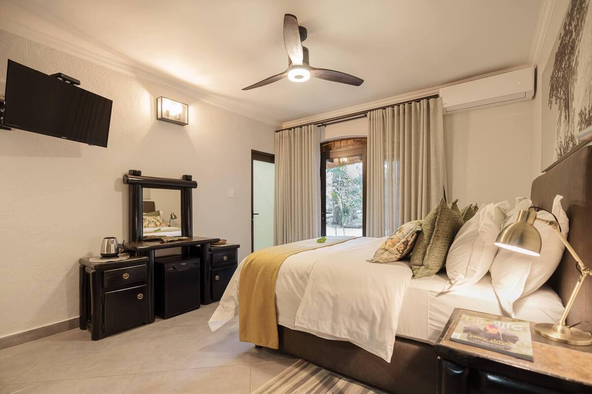 A bedroom with a bed, bedside table, lamp, TV, ceiling fan, and a view of the outdoors.