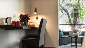 A modern room with a dark grey upholstered chair, a small table with a lamp, a shelf with kitchen items, and a view of a tree through a window. - Kruger Eden Lodge (Nkomazi)