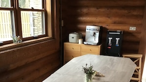 Une longue table en bois sur laquelle se trouvent un vase de fleurs, un bloc-notes et une clé. Sur un meuble en bois, on aperçoit une machine à café et un distributeur d’eau.