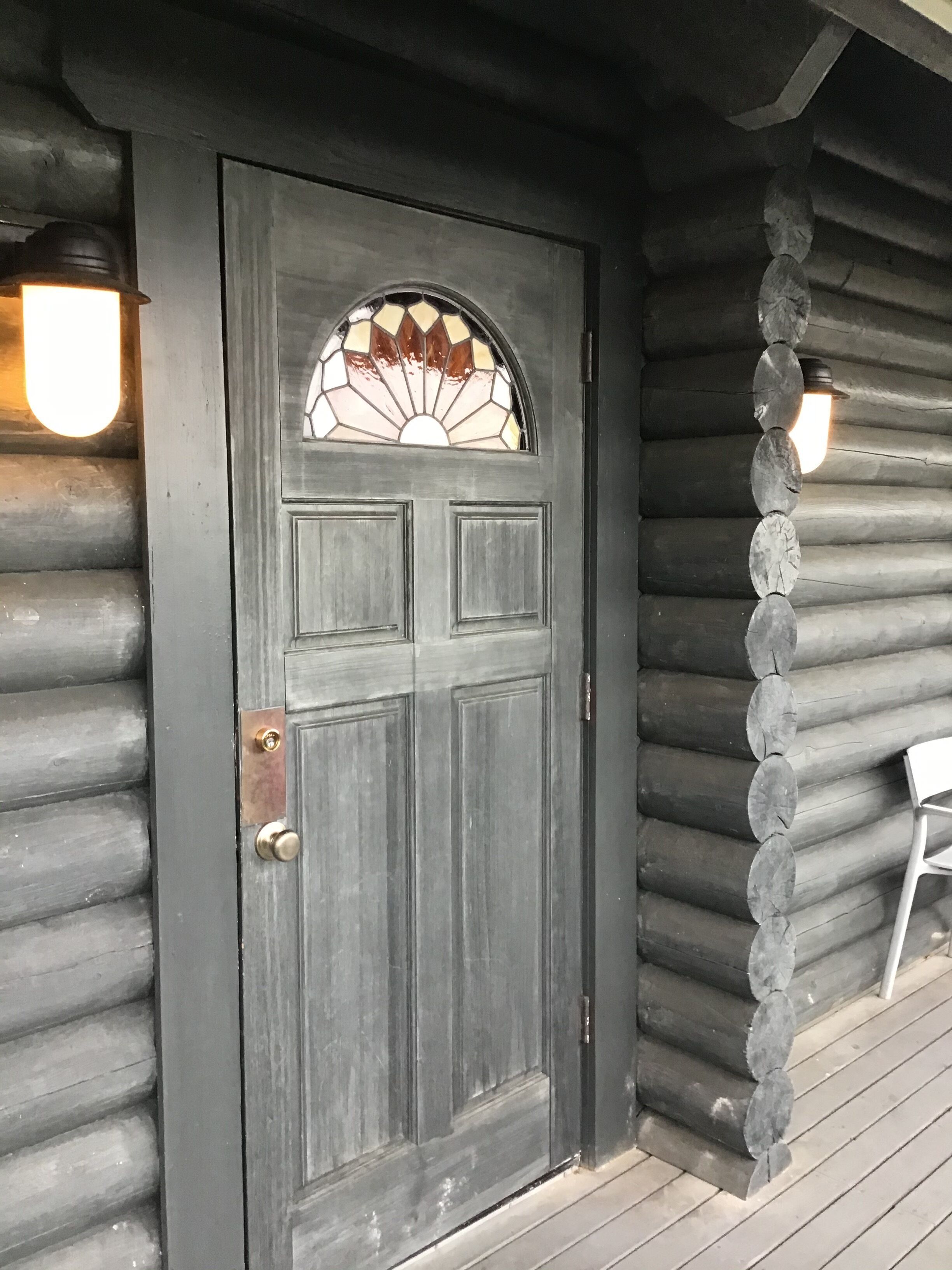 A log cabin with a dark wooden door, a stained glass window, and two wall-mounted lights.