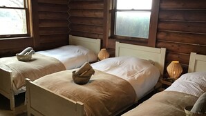 Une pièce avec trois lits en bois, chacun doté d’un matelas blanc et d’une couverture beige. On trouve des paniers en osier sur les lits, ainsi qu’une lampe sur la table de chevet à côté de chaque lit.