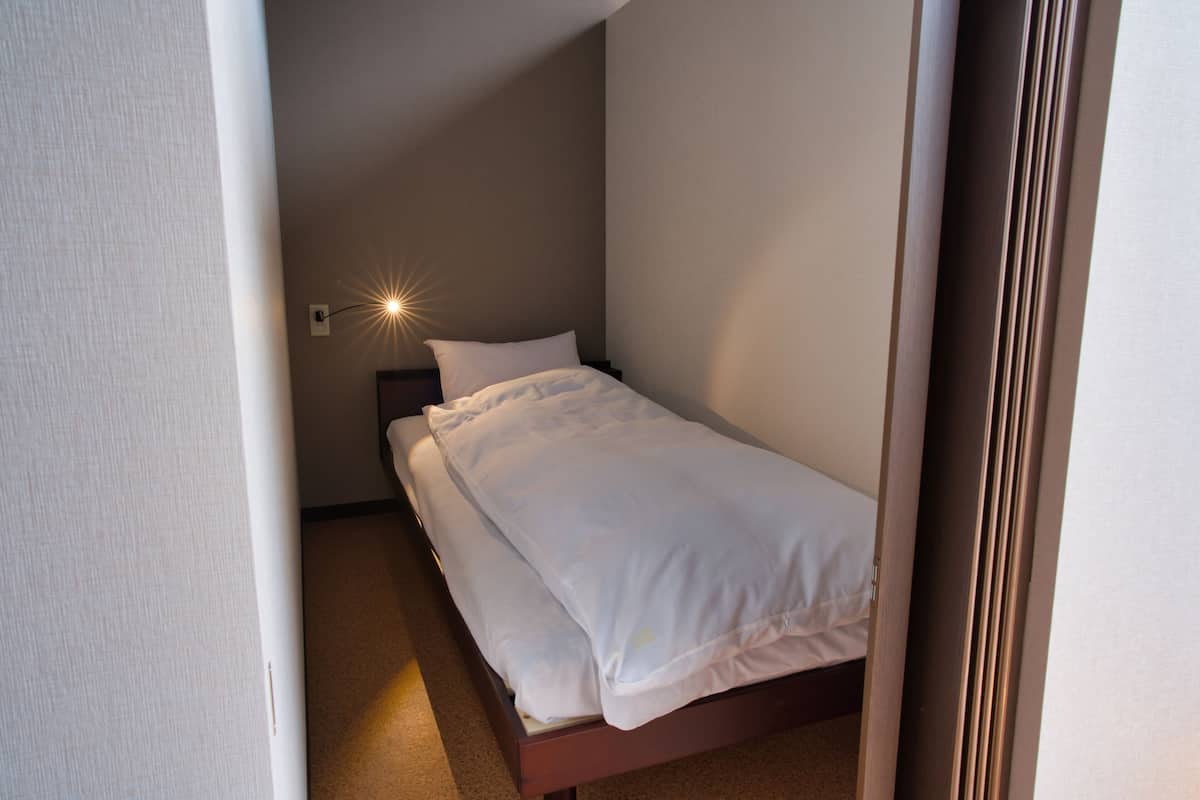 Een klein, netjes opgemaakt bed in een compacte kamer met één wandlamp.