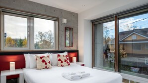 Moderni hotellihuone, jossa on suuri sänky, kaksi yöpöytää, yölamppu, ikkuna, josta on näkymä ulos, ja punaisella kehyksellä varustettu peili seinällä.