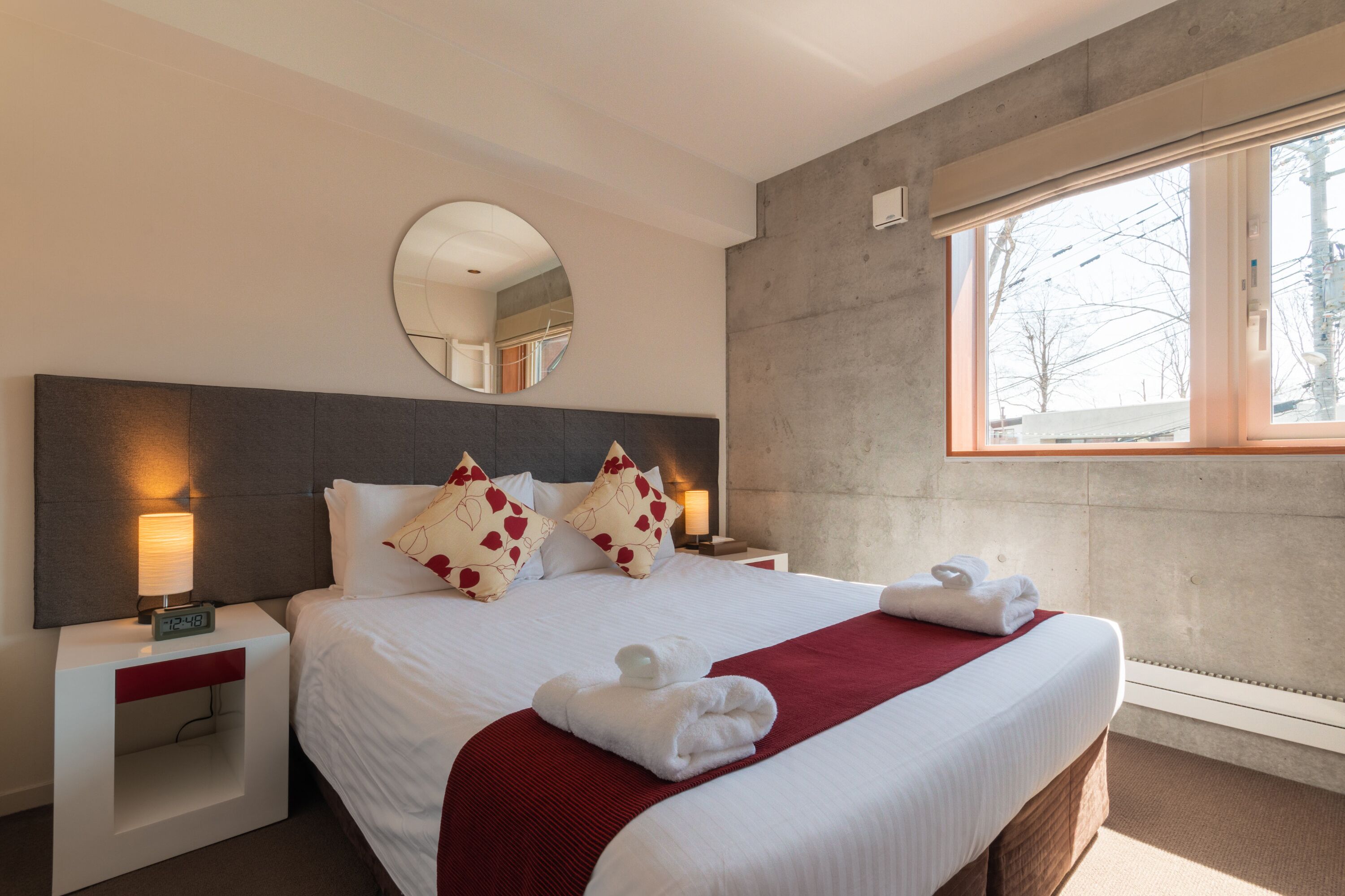 Habitación de hotel con una cama grande, un espejo redondo, una mesita de noche con lámpara y una ventana con vistas a árboles.