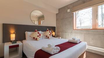 Habitación de hotel con una cama grande, un espejo redondo, una mesita de noche con lámpara y una ventana con vistas a árboles.
