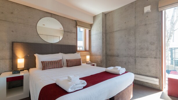 A modern hotel room with a large bed, a round mirror, a nightstand with a clock, and a window providing natural light. - Yuki Ten by H2 Life (Kutchan)