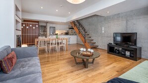 Un salon moderne avec un canapé gris, une table basse en bois sur laquelle sont posés des pièces d’échecs, un téléviseur sur un meuble noir et un escalier menant à la cuisine.