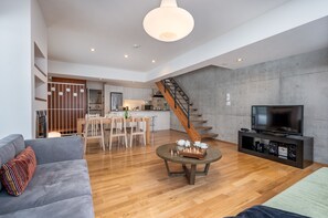 A modern living room with a grey sofa, a wooden coffee table with chess pieces, a TV on a black stand, and a staircase leading to a kitchen.