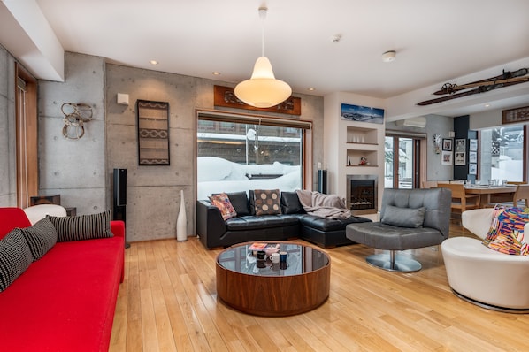 Una sala de estar moderna con un ventanal grande, una chimenea y diversas opciones de asientos, incluyendo un sofá rojo, un sofá modular negro y un sillón gris.