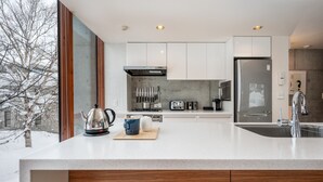 A modern kitchen with white cabinets, stainless steel appliances, and a large window overlooking a snowy landscape. - Kitanishi 3 by H2 Life (Kutchan)