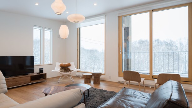 Ein modernes Wohnzimmer mit einem Flachbildfernseher, Ledersofas, einem Couchtisch und großen Fenstern mit Blick auf Bäume.