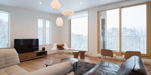 Een moderne woonkamer met een flatscreen televisie, leren banken, een salontafel en grote ramen met uitzicht op bomen.