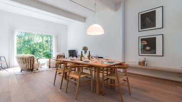 A dining area with a wooden table set for six, surrounded by chairs, with a large window providing a view of greenery outside.
