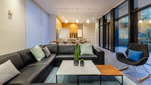 A modern living room with a sectional sofa, a coffee table, and a rocking chair. There is a dining area in the background with a kitchen visible.