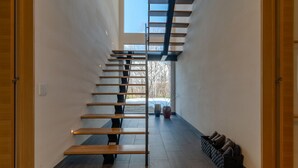 A modern interior with a central staircase, a shoe rack, and a view through a window.