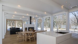 A modern kitchen with a large island, wooden dining table, and chairs. There is a fireplace and large windows offering views of a snowy landscape.