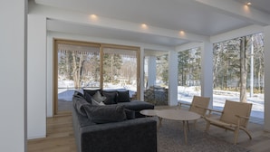 A modern living room with a grey sofa, a round coffee table, and wicker chairs. Large windows offer a view of a snowy outdoor area.