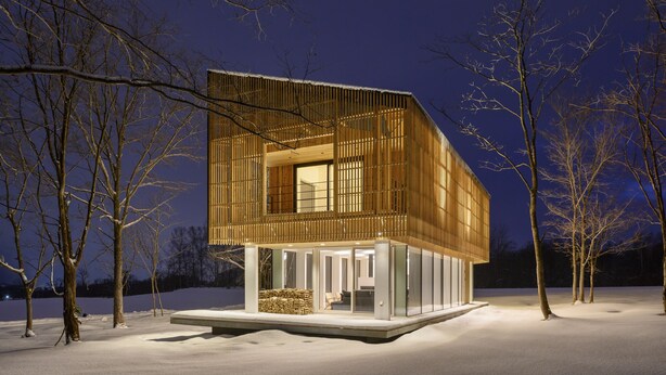 A modern, wooden cabin with a glass entrance, surrounded by snow-covered ground and bare trees.