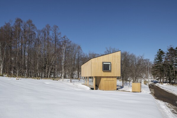 Una cabaña de madera moderna con un ventanal pequeño, rodeada por un paisaje nevado y un bosque.