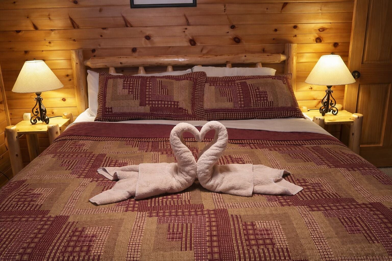 A wooden cabin-style room with a bed, two nightstands with lamps, and a heart-shaped towel arrangement on the bed.