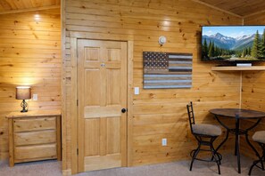 Ein Holzhütten-Interieur mit Holztür, einem an der Wand montierten Fernseher, der eine Berglandschaft zeigt, einem kleinen Tisch mit zwei Stühlen und einer Lampe auf einem Nachttisch.