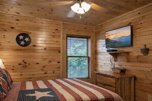 Ein Holzhütten-Zimmer mit einem Bett, einem an der Wand montierten Fernseher und einem Fenster mit Jalousien.
