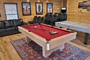 Ein Billardtisch mit roter Filzoberfläche, eine Spielstange und Billardkugeln, umgeben von schwarzen Sesseln und einer Holzwand mit gerahmten Filmplakaten.