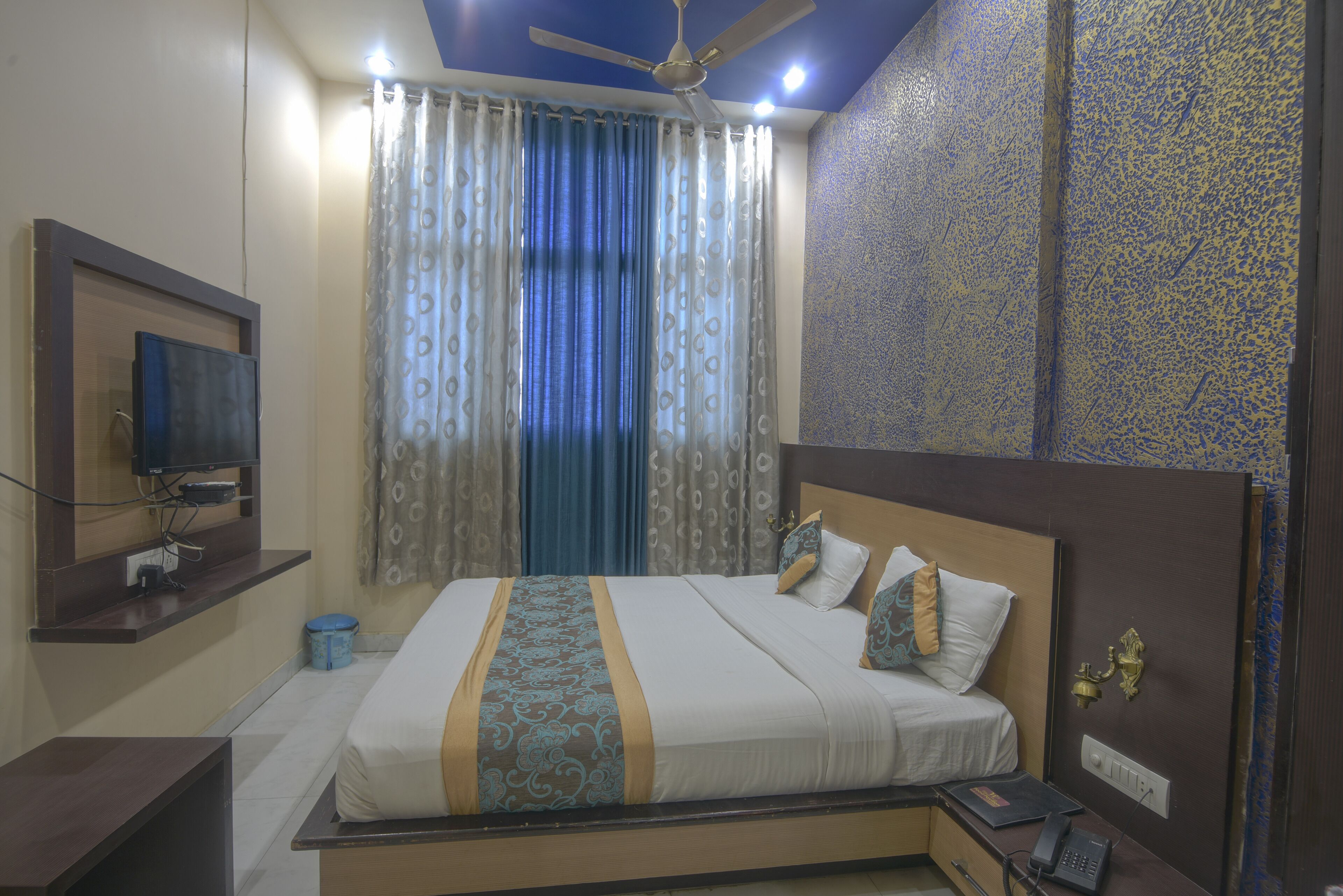A hotel room with a bed, a TV mounted on the wall, a ceiling fan, and a patterned wall.