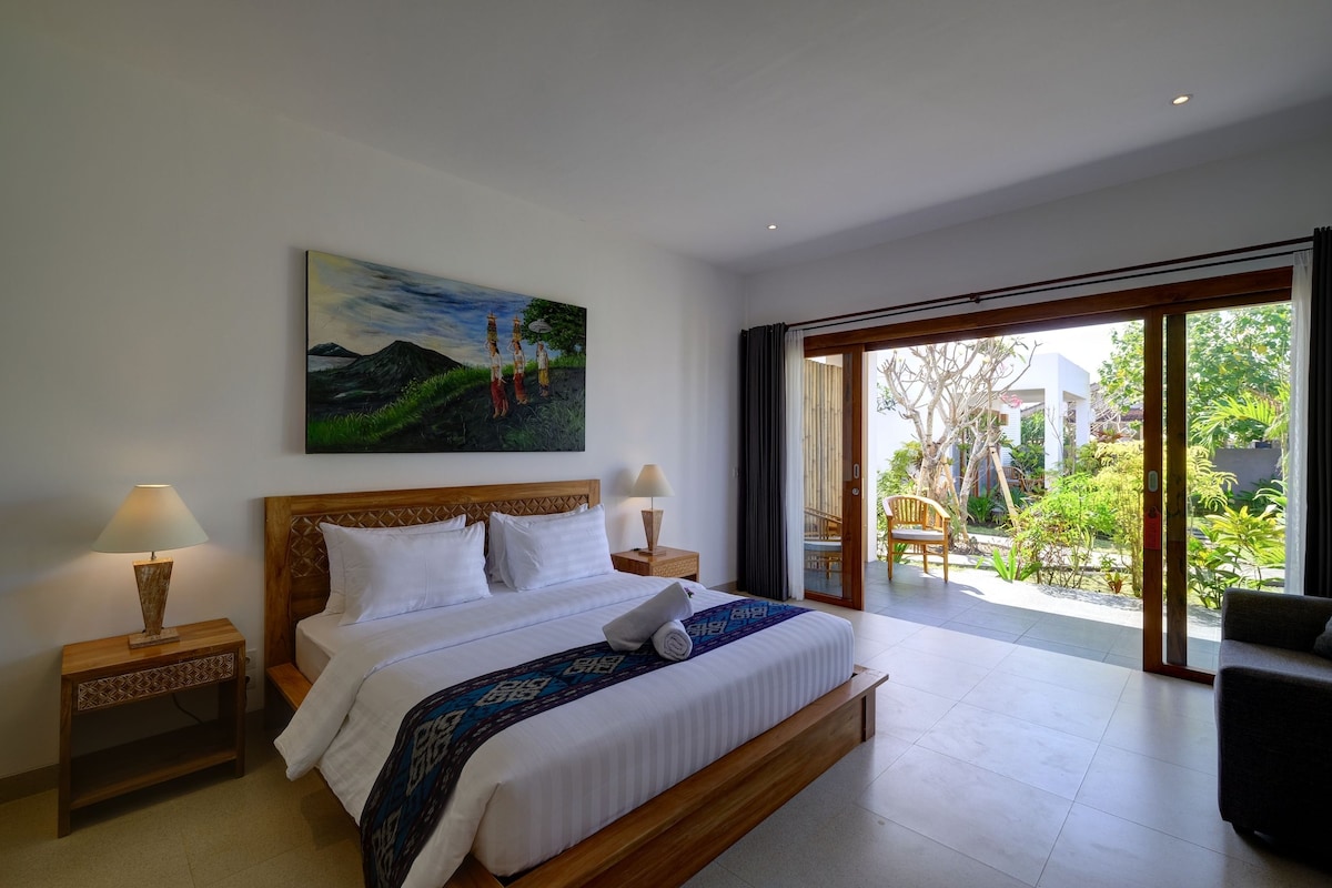 Ein Hotelzimmer mit einem großen Bett, zwei Nachttischen, einem Bild an der Wand und Blick auf den Garten durch die Glastür.